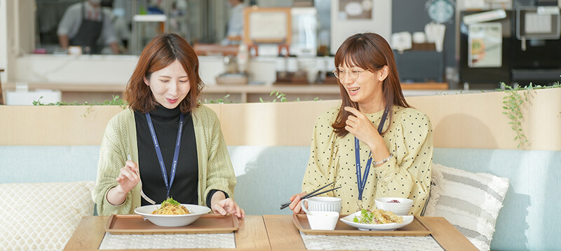 明るいカフェのような社内食堂で、パスタや和定食のランチを楽しみながら、にこやかに談笑する2人の女性社員。