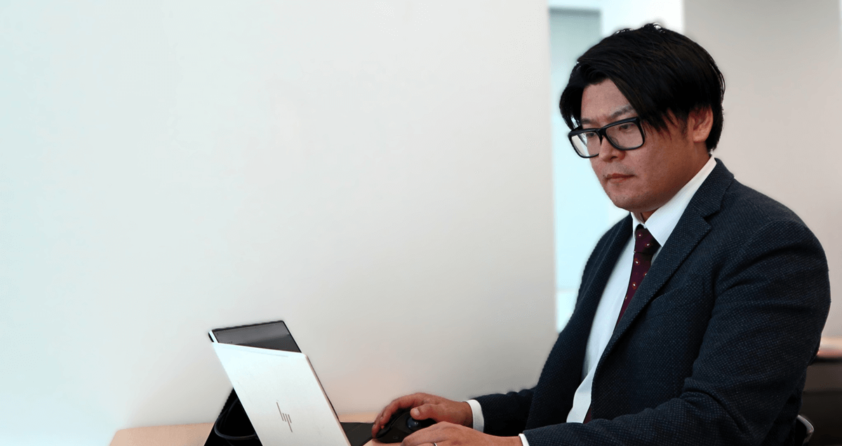 眼鏡をかけ、スーツを着た男性が、ノートパソコンの画面を真剣な表情で見つめています。