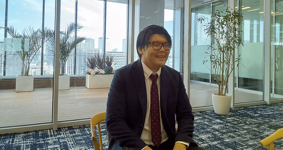 ガラス張りの開放的なオフィスで、スーツ姿の男性が椅子に座って談笑している。背景には観葉植物や都会のビル群が見える。