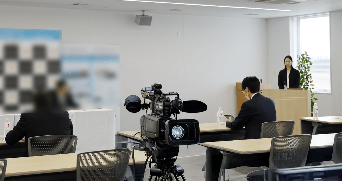 イベント会場の設営風景。前方には壇と話者がおり、中央には撮影用のビデオカメラ、手前には参加者が座れる机と椅子が並んでいます。