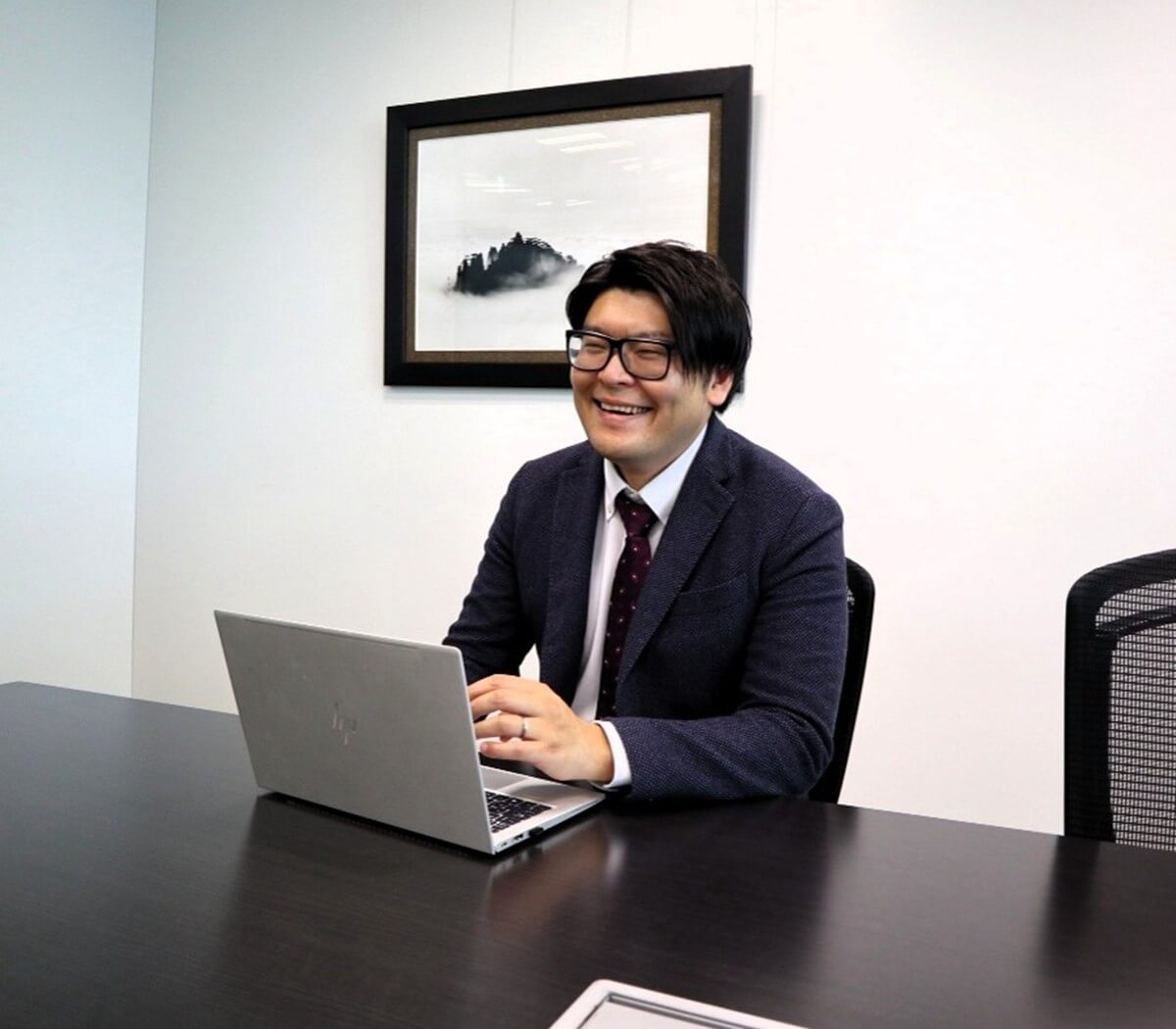 会議室で、ノートパソコンの前で笑顔を見せる眼鏡をかけた男性。