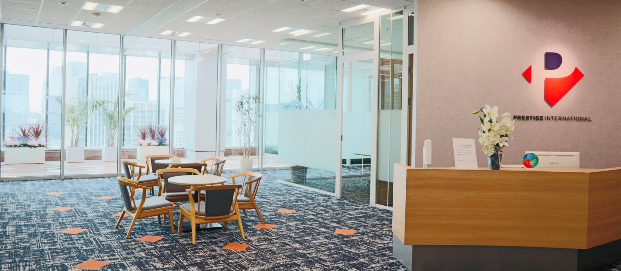 明るく開放的なオフィスのエントランスラウンジ。受付カウンターの横には打ち合わせスペースがあり、大きな窓の外にはバルコニーと都会のビル群が見える。