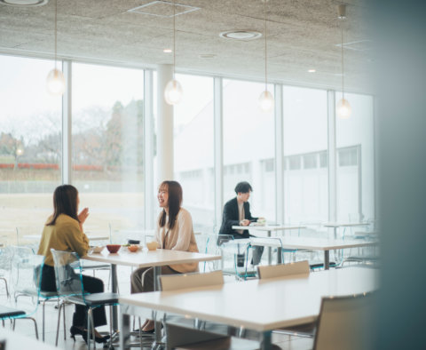 大きな窓から自然光が差し込む明るく開放的なカフェテリアで、テーブルを囲んで楽しそうに談笑している2人の女性と、奥の席で食事をしている男性。