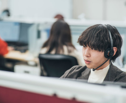 オフィスでヘッドセットを装着し、真剣な表情で電話対応業務を行っている男性。背景には他のデスクで仕事をしているスタッフの姿もぼやけて見えている。