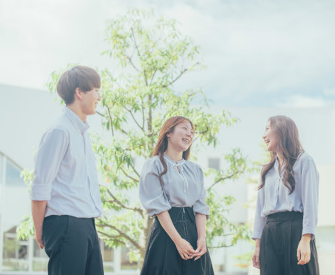 屋外の白い建物の前にある木の下で、爽やかな服装の若い男性1人と女性2人が、笑顔でリラックスして会話を楽しんでいる様子。