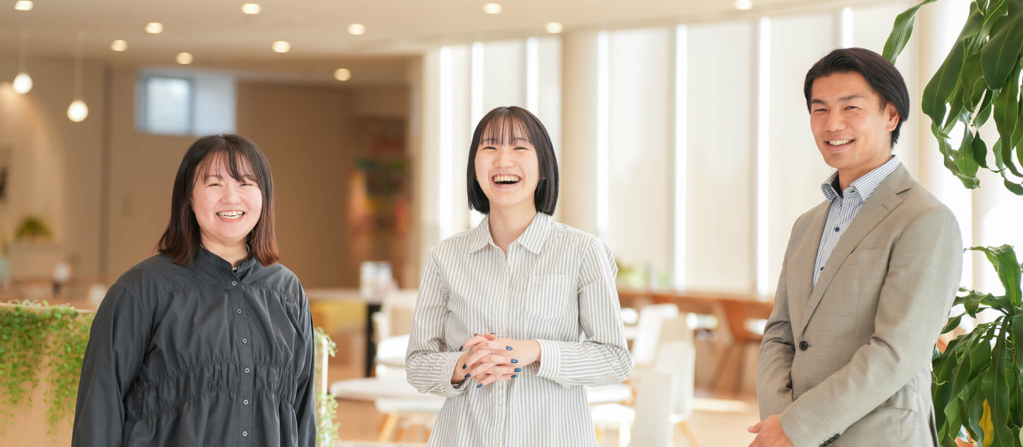 明るく開放的なオフィスで、笑顔で並ぶ3人の男女。左側に黒いシャツの女性、中央にストライプ柄のシャツの女性、右側にグレーのスーツを着た男性が、和やかな雰囲気で正面を向いている様子。