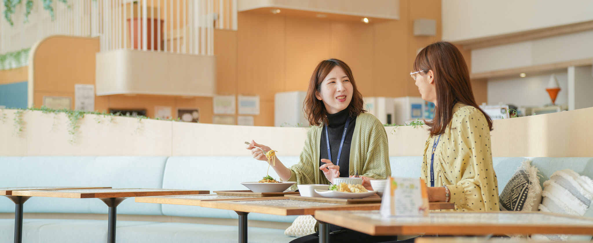 モダンで清潔感のあるカフェスペースのソファ席で、パスタなどのランチを食べながら楽しそうに談笑する2人の女性。背景には木目調のインテリアと明るい光が差し込む窓がある。