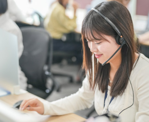 オフィスでヘッドセットを装着し、パソコンに向かって優しく微笑みながら業務を行う女性。清潔感のある白いブラウスを着用している。