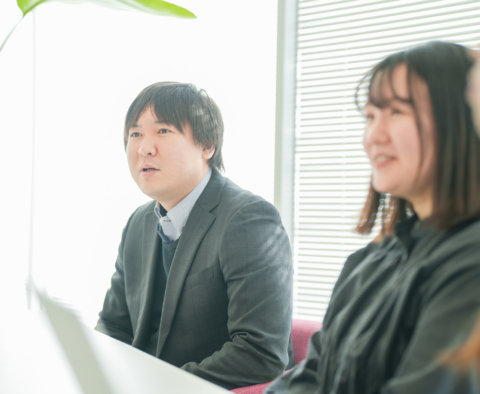 明るい会議スペースで、真剣な表情で意見を述べるスーツ姿の男性社員。隣には話を聞く女性社員が座っている。