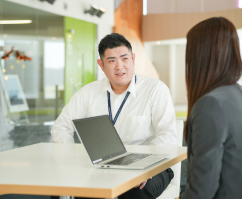 オープンな打ち合わせスペースで、ノートパソコンを開きながら対面の相手と相談する白いシャツの男性。背景にはアクセントカラーの黄緑色の壁が見える。