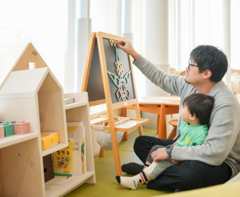 社内のキッズスペースで、黒板にマグネットのおもちゃを貼り付けて子供と一緒に遊ぶ男性社員。家庭的な温かさを感じる風景。
