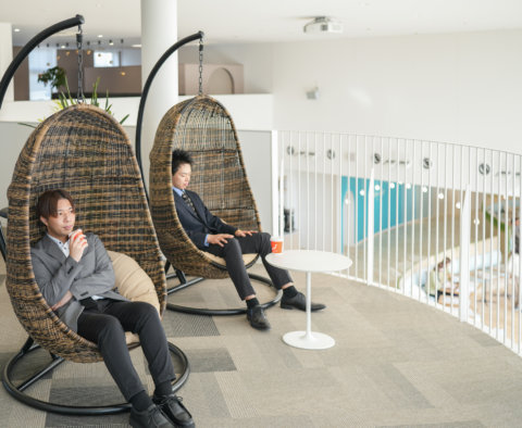 オフィスの開放的なスペースにある卵型のハンギングチェアに座り、コーヒーを飲みながらリラックスする2人の男性。