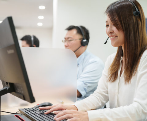 コールセンターの業務スペースで、ヘッドセットをつけてタイピングをしながら笑顔を見せる女性。横顔のアップ。