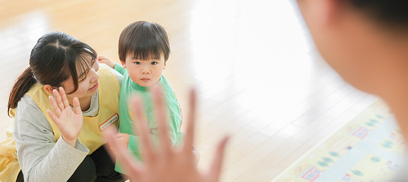 保育施設で、黄色のエプロンを着た保育士の女性が、床に座って緑色の服を着た小さな男の子に優しく話しかけている様子。