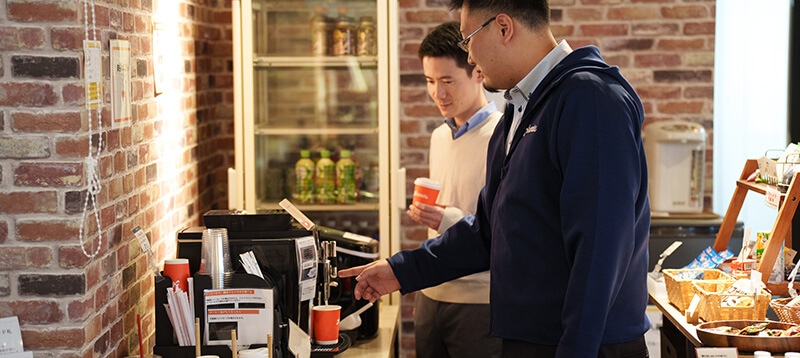 レンガ調の壁がおしゃれな休憩スペースで、本格的なコーヒーマシンを操作する男性と、コーヒーを待つもう一人の男性社員。