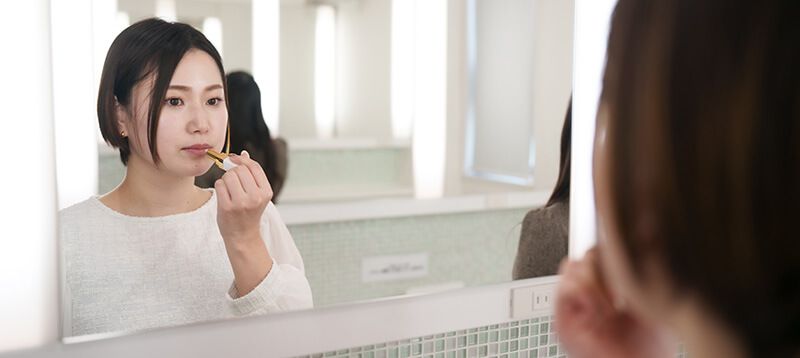 清潔感のある白いパウダールームの鏡の前で、リップを塗り直して身だしなみを整える白いブラウス姿の女性。