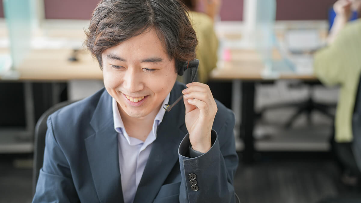 コールセンターで業務中の男性社員。ヘッドセットに手を添え、パソコンの画面を見ながら満足そうに口角を上げているアップの写真。