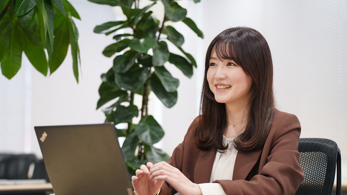 オフィスで茶色のジャケットを羽織り、ノートパソコンを前にして明るく微笑む女性社員。背景には大きな観葉植物があり、ナチュラルで落ち着いた雰囲気。