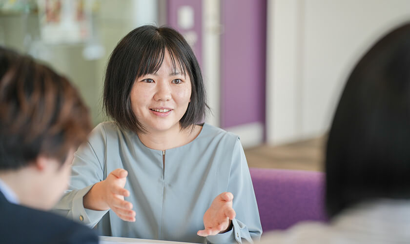 打ち合わせ中、水色のトップスを着た女性が身振り手振りを交えて楽しそうに話している様子。前向きで活気のあるコミュニケーションシーン。