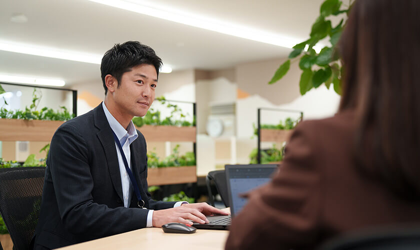 整えられたオフィスデスクでノートパソコンを操作しながら、対面の相手の話を真剣かつ穏やかな表情で聞くスーツ姿の男性社員。