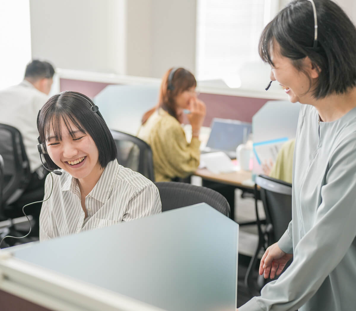 活気あるコールセンターのオフィス風景。手前でヘッドセットをつけた女性社員が楽しそうに笑い、その隣で別の女性社員が笑顔で話しかけている。