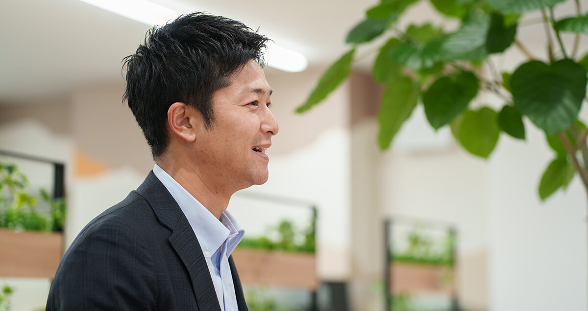 日差しが差し込む明るいオフィスで、観葉植物を背景に爽やかな笑顔を見せるスーツ姿の男性社員の横顔。