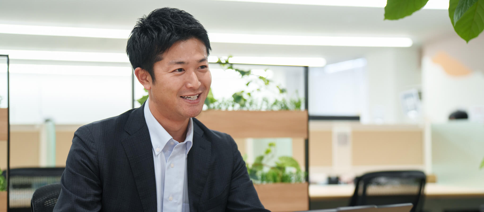 オフィスのデスクで、親しみやすく自信に満ちた笑顔を浮かべるスーツ姿の男性社員。信頼感を感じさせる上半身のアップショット。