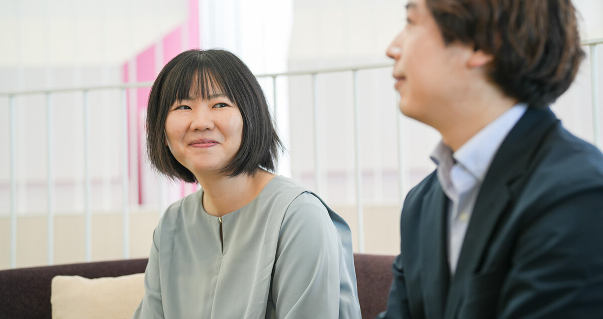 打ち合わせ中、隣に座る社員の話に耳を傾け、優しく微笑む水色のブラウスを着た女性社員。自然なコミュニケーションの様子。