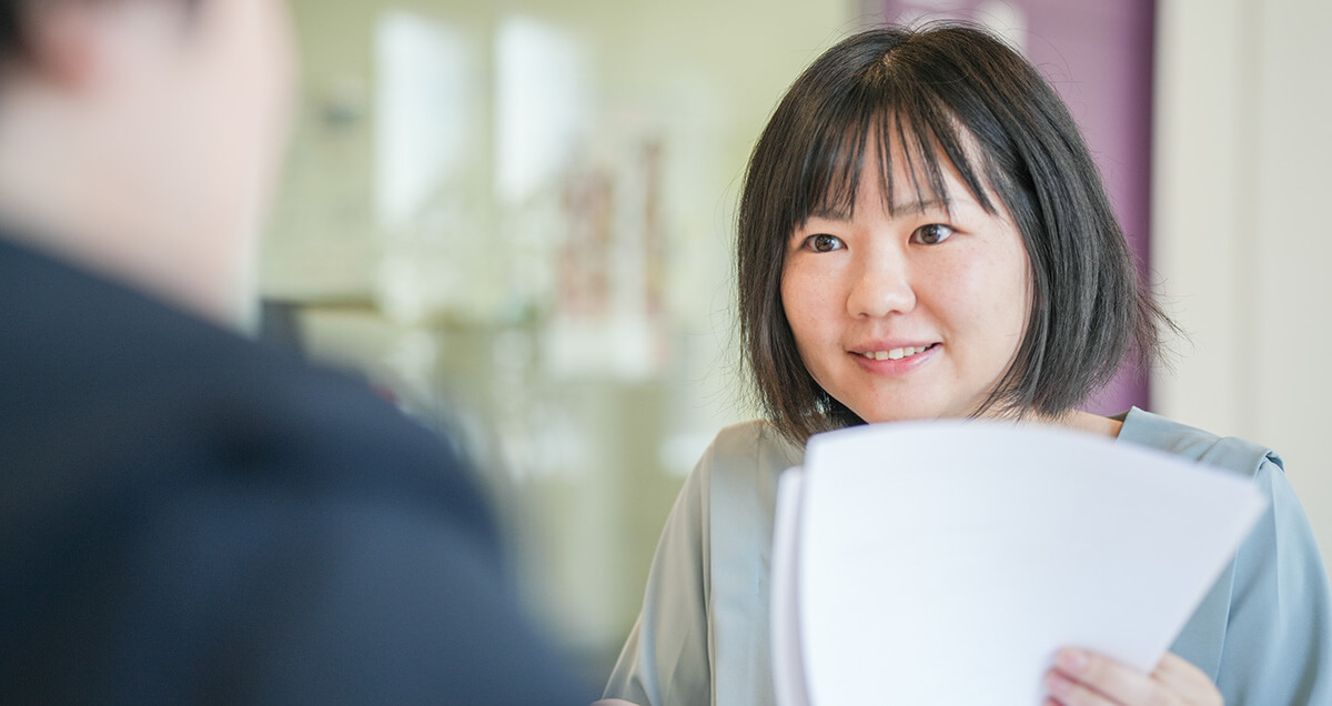 数枚の資料を手に持ちながら、明るい表情で打ち合わせに参加する女性社員。活気あるオフィスの雰囲気を伝えるカット。