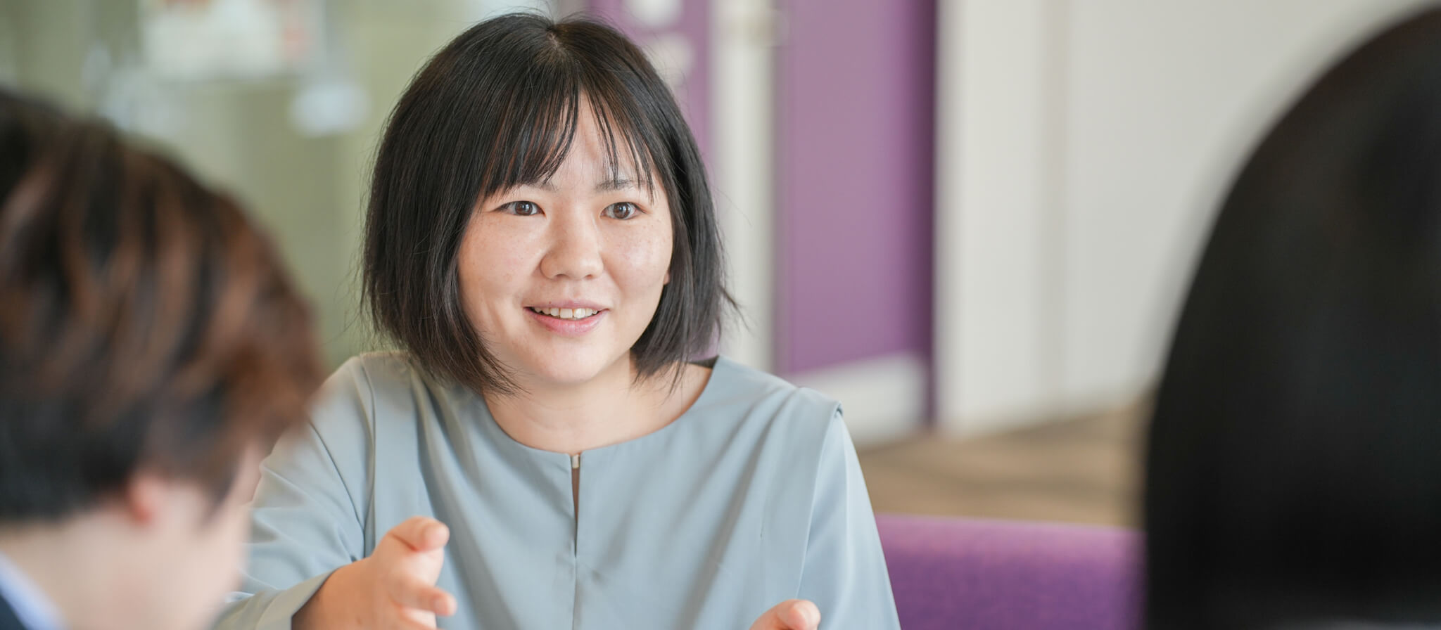 明るく柔らかな表情で、対話の相手を見つめて微笑むショートヘアの女性社員。清潔感のある水色のトップスを着用している。